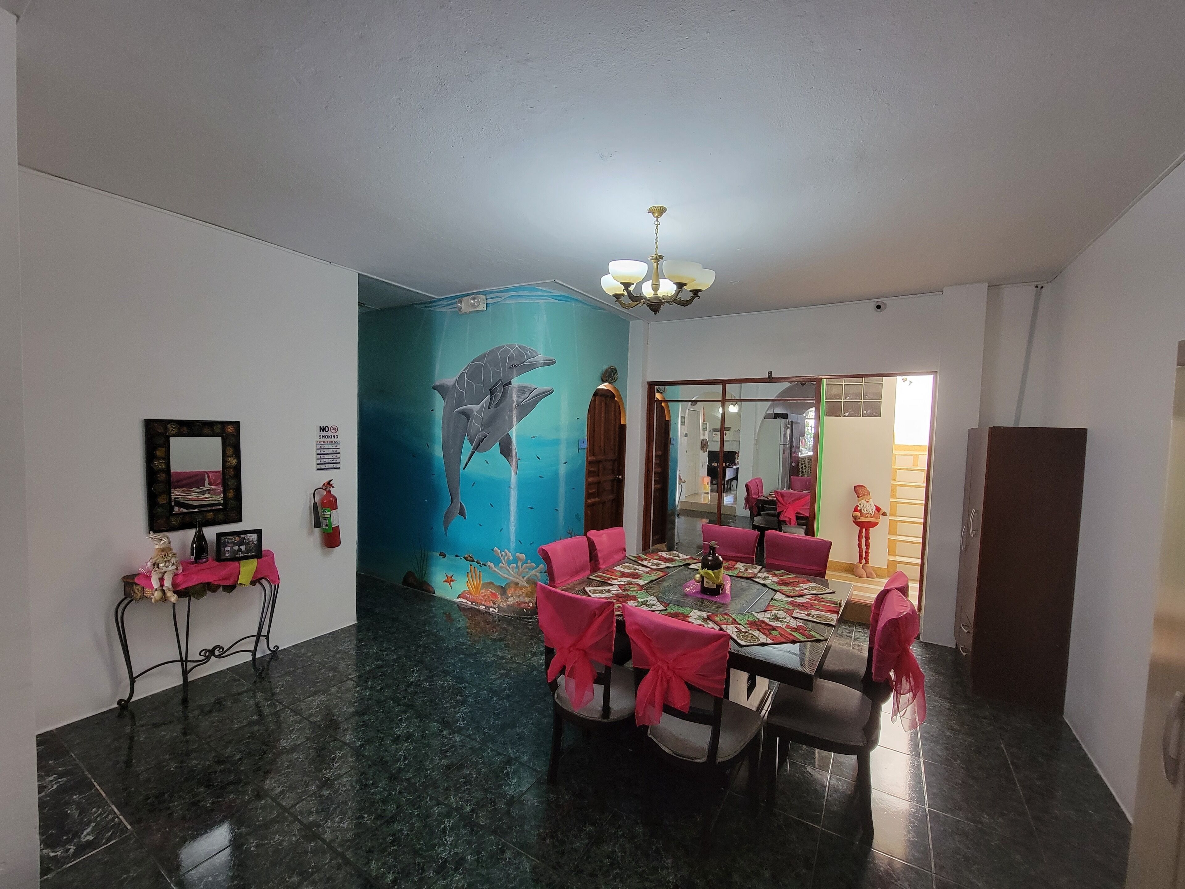 A dining area with a table set for six, chairs with pink covers, a wall mural of a dolphin, and a chandelier.