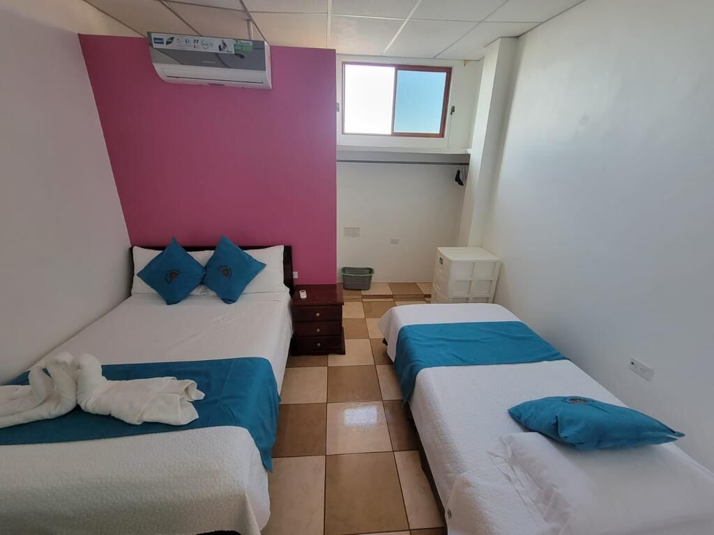 A hotel room with two beds, a window, an air conditioning unit, a small table, and a trash bin.