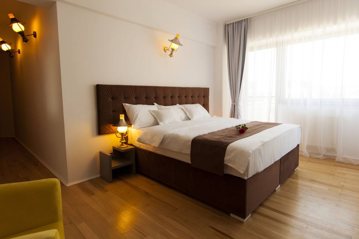Ein ordentlich eingerichtetes Schlafzimmer mit einem großen Bett, einem Nachttisch mit Lampe und einem Fenster mit Vorhängen.