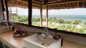 A bathroom with a white sink, a window with a view of the ocean and a beach, and a thatched roof structure. - Jua Retreat (Michamvi)