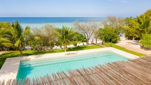 A clear blue pool with a wooden deck, surrounded by lush greenery and palm trees, overlooking a beach and ocean. - Jua Retreat (Michamvi)