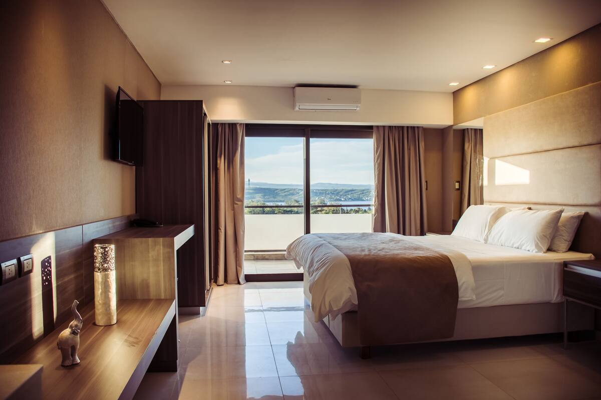 A modern hotel room with a large bed, a flat-screen TV, a desk, and a view of the outdoors.