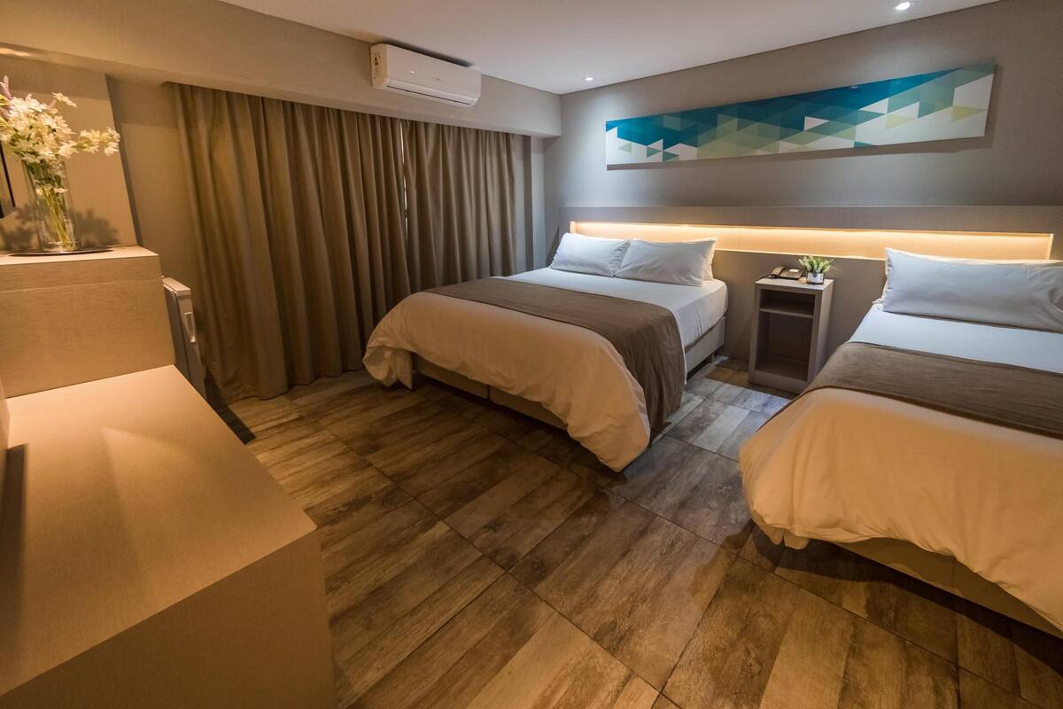 A hotel room with two beds, a desk, a chair, a vase with flowers, a wall-mounted air conditioner, and a geometric wall art piece.
