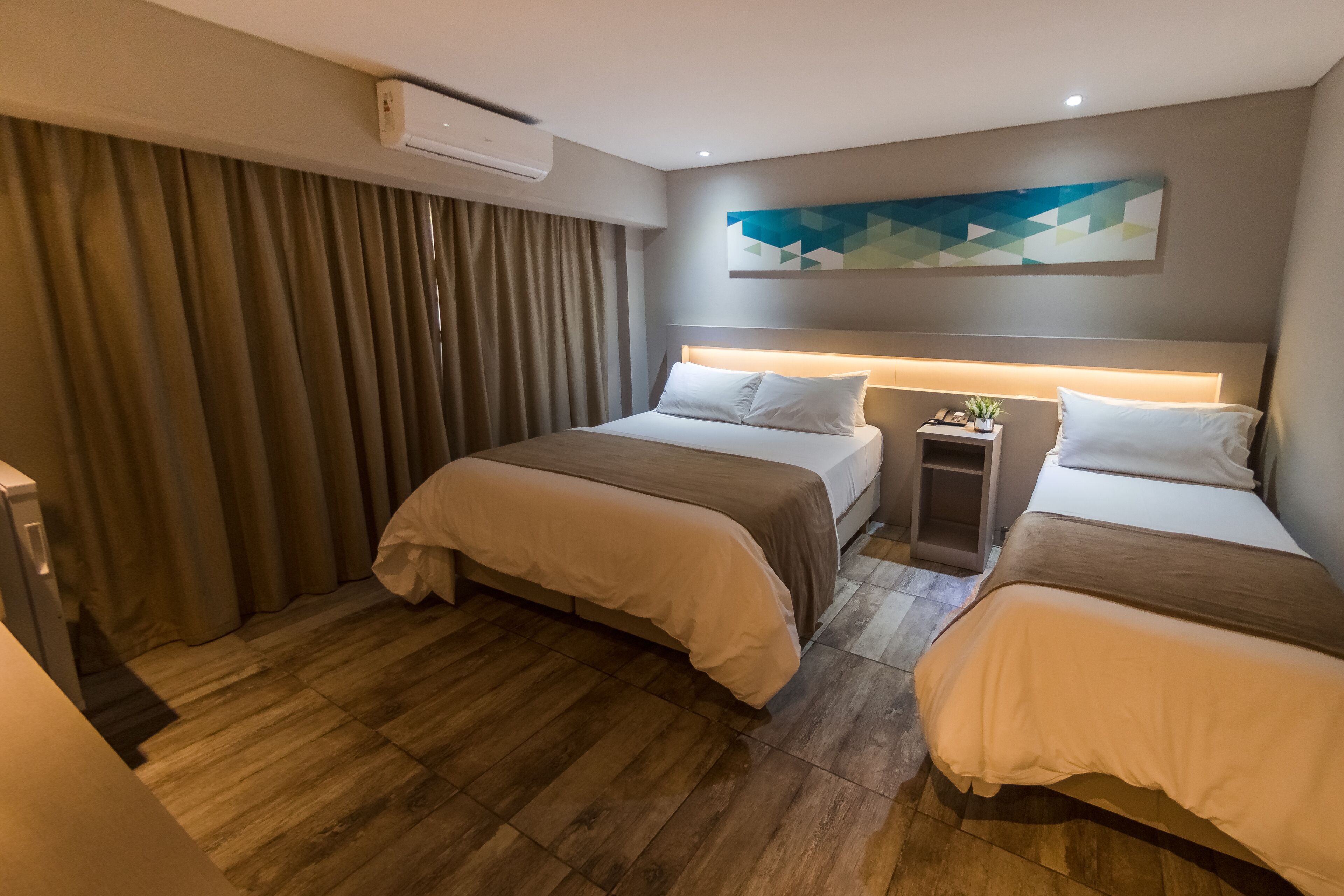A hotel room with two beds, a bedside table with a lamp, a wall-mounted air conditioner, and a geometric wall art piece.