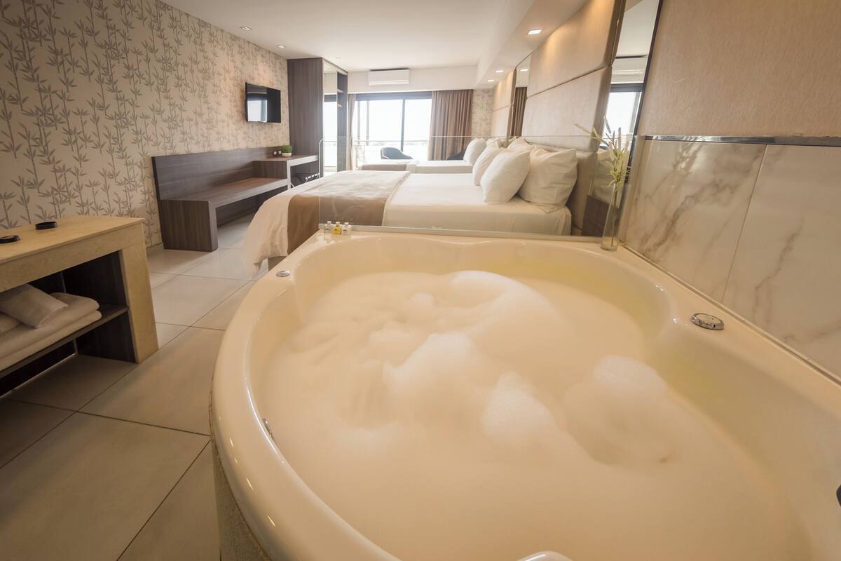 A hotel room with a large bathtub, a bed, a desk, and a mirror.