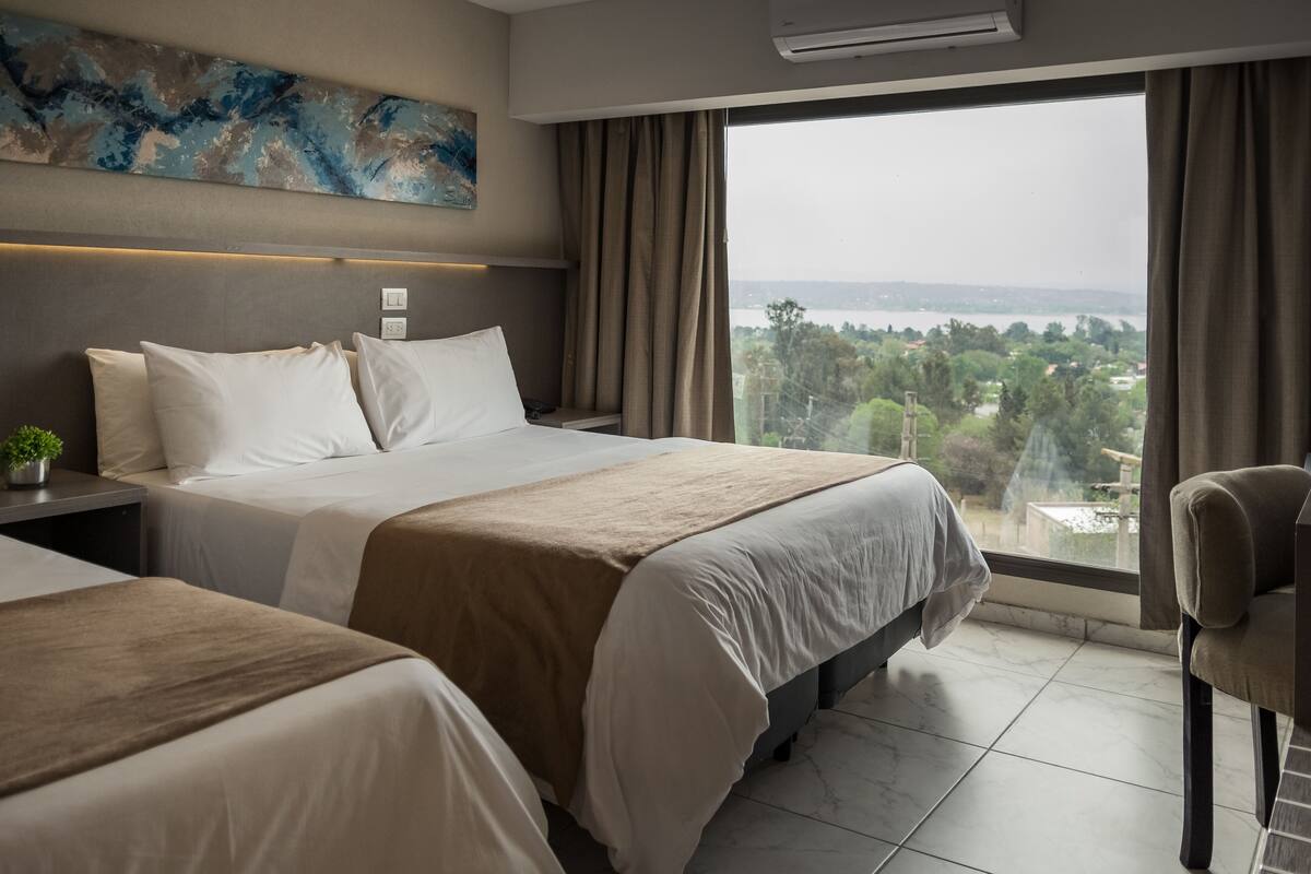 A hotel room with two beds, a large window with a view, a painting on the wall, and a plant on the nightstand.