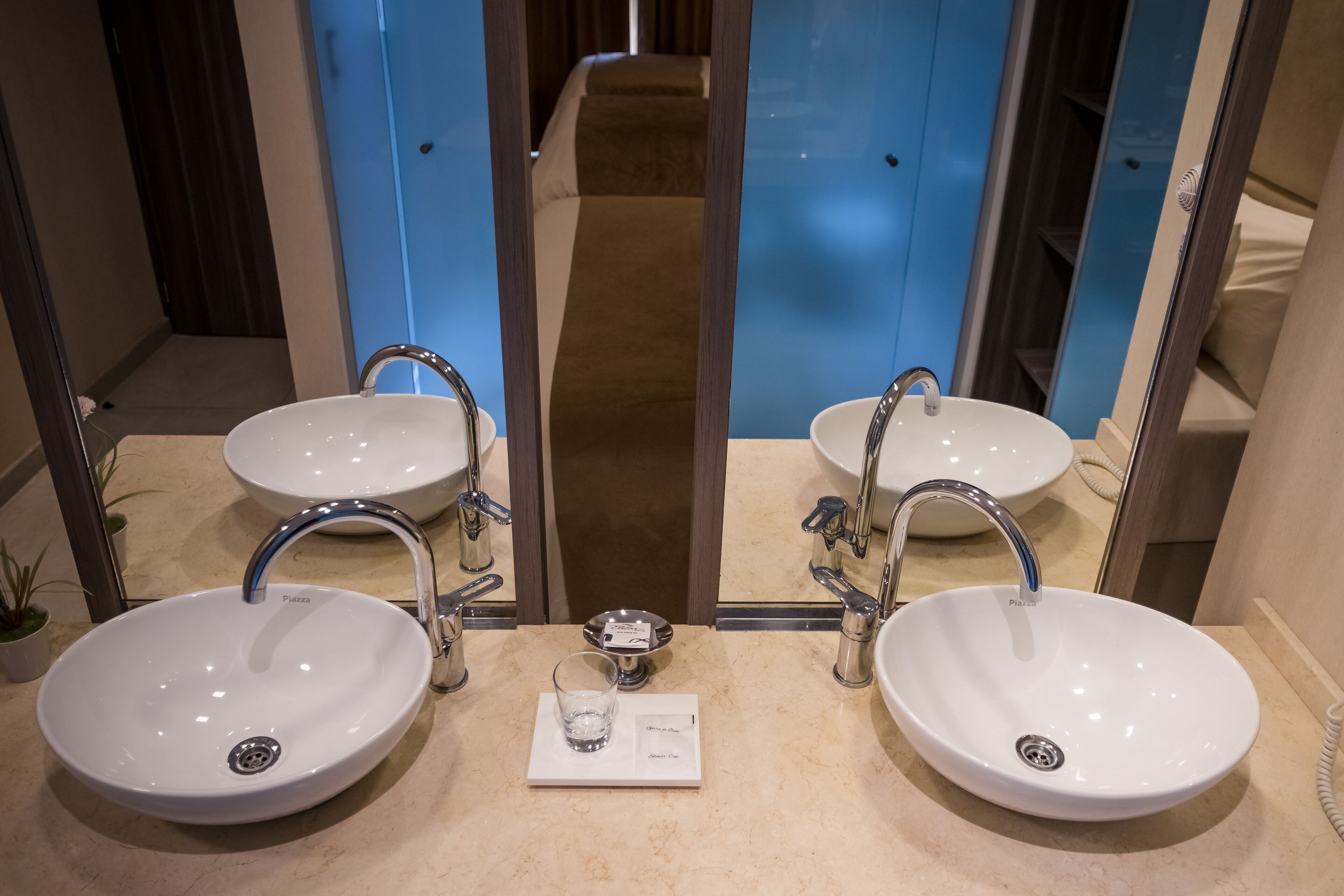 Un baño con dos lavamanos blancos, cada uno con un espejo encima. Hay una balanza entre los lavamanos y un vaso pequeño con agua sobre la balanza.