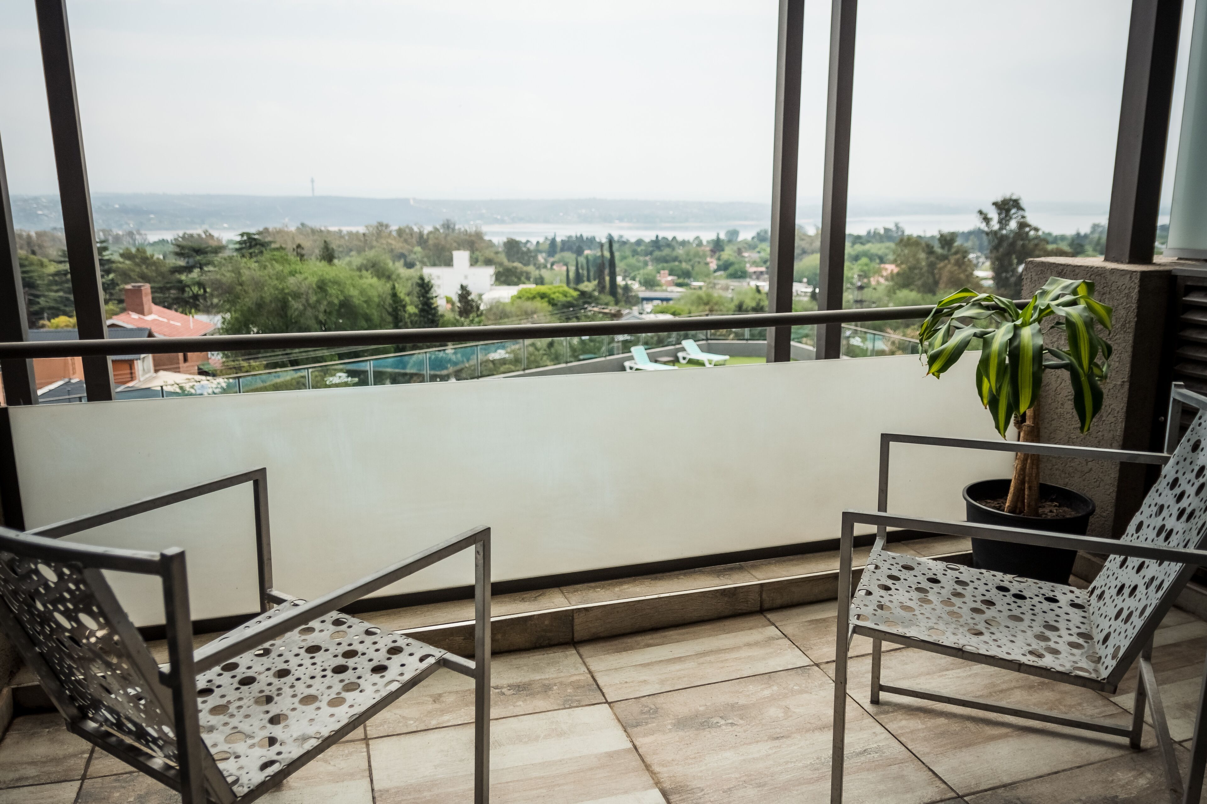 Een balkon met twee metalen stoelen, een bloempot en uitzicht op een stadsgezicht.