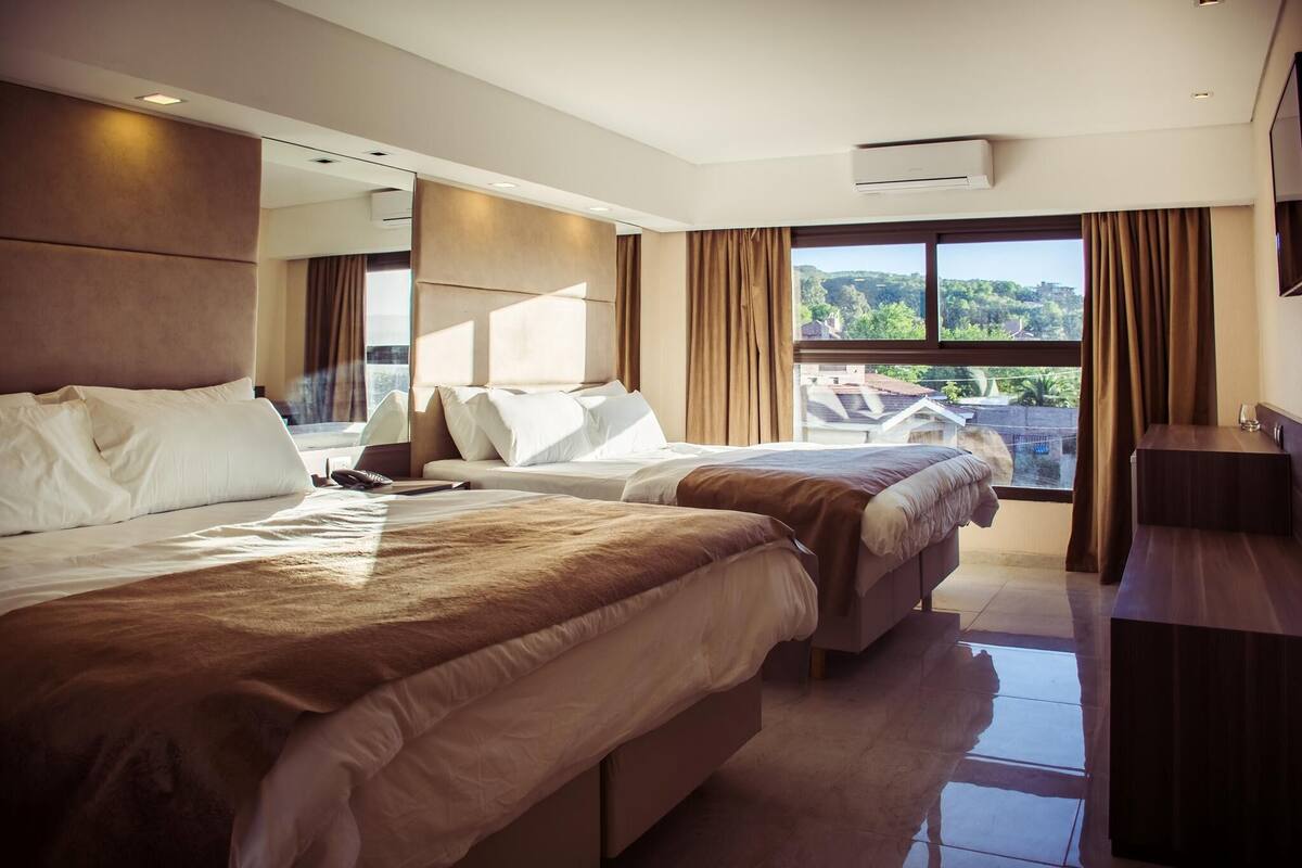 A hotel room with two beds, a desk, a large window with curtains, and an air conditioning unit.