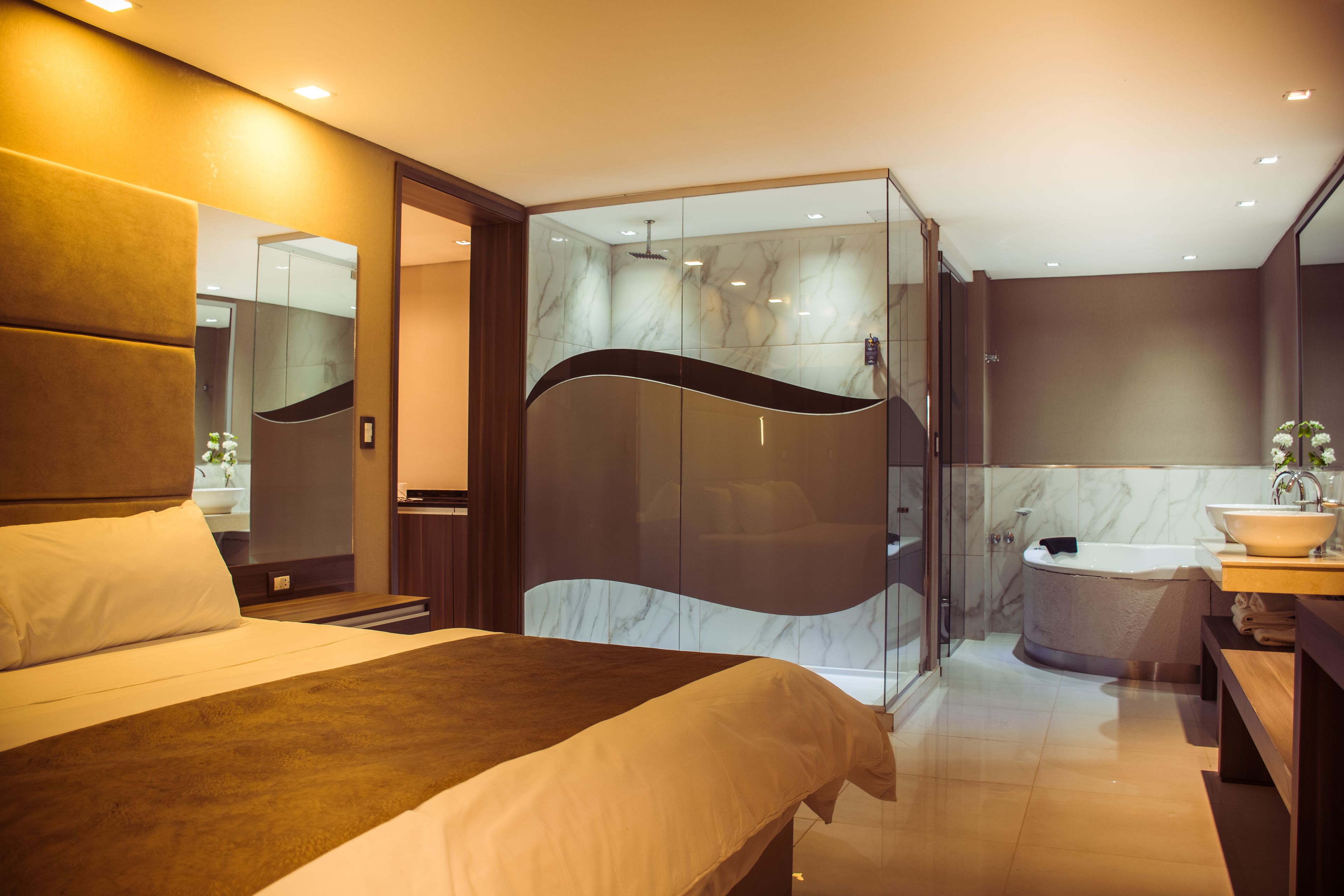 Una habitación de hotel moderna con una cama grande, un cabina de ducha con paredes de cristal, un baño con suelo de mármol y un banco de madera.