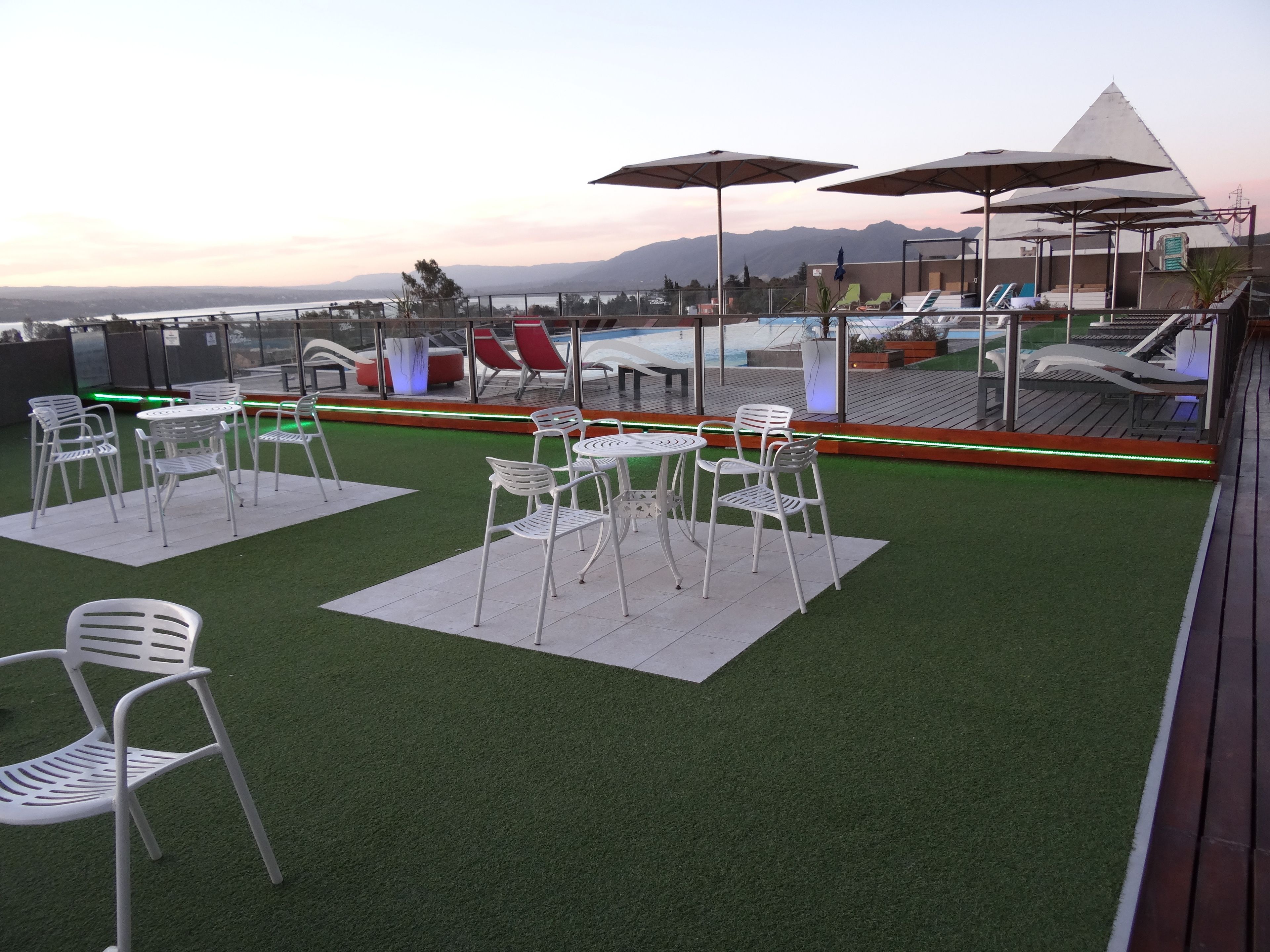 A rooftop terrace with artificial grass, white metal chairs, and umbrellas, overlooking a pool and mountains.