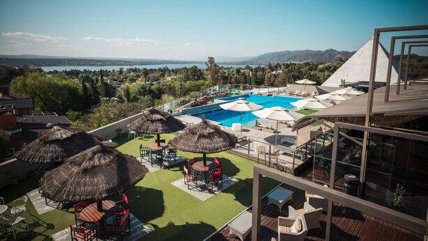 Una piscina en la azotea con vistas a un lago y montañas, rodeada de asientos exteriores y sombrillas.