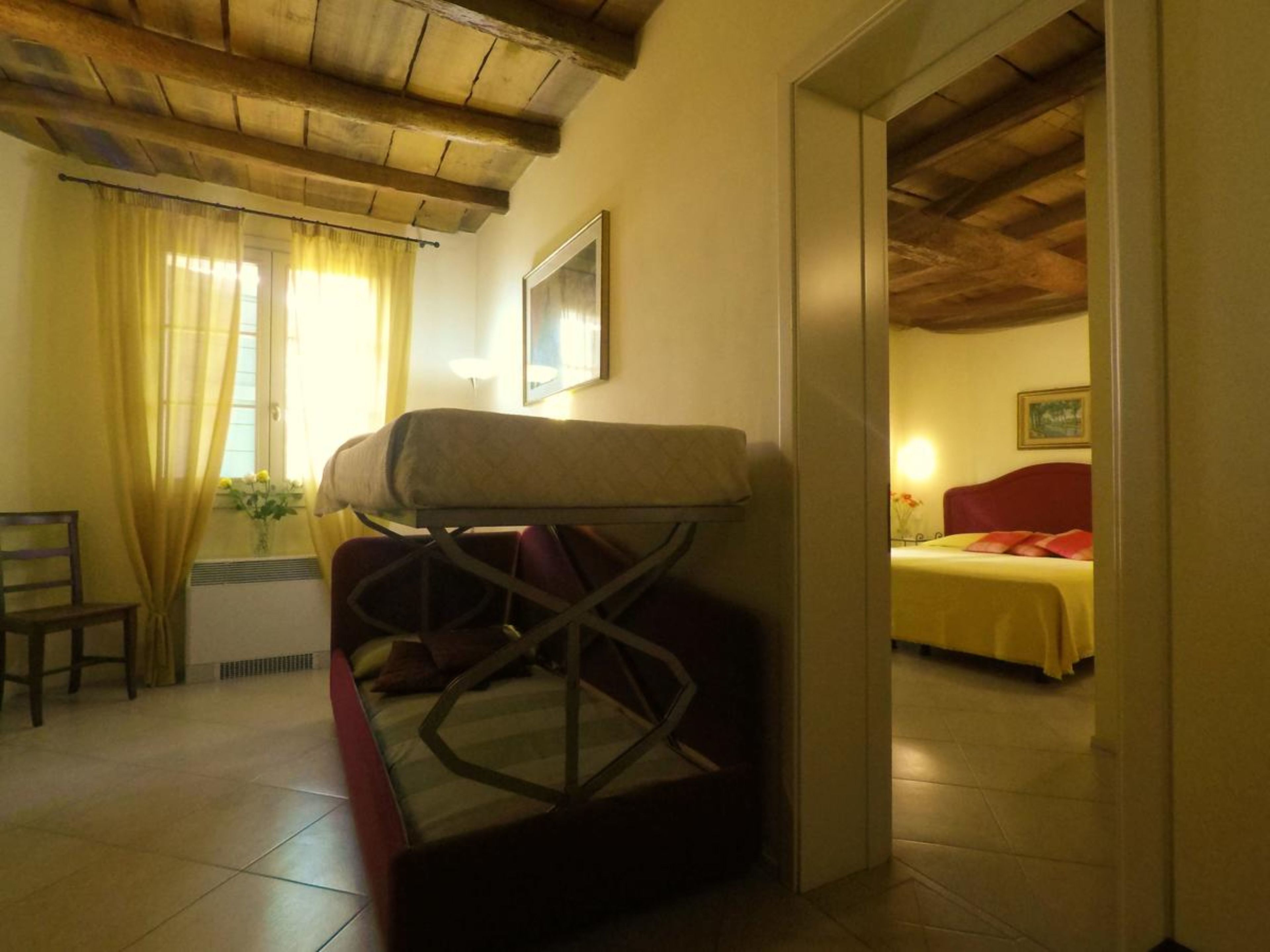 Una habitación con una cama, una silla y una mesa. Hay una ventana con cortinas, un cuadro en la pared y un techo de madera.