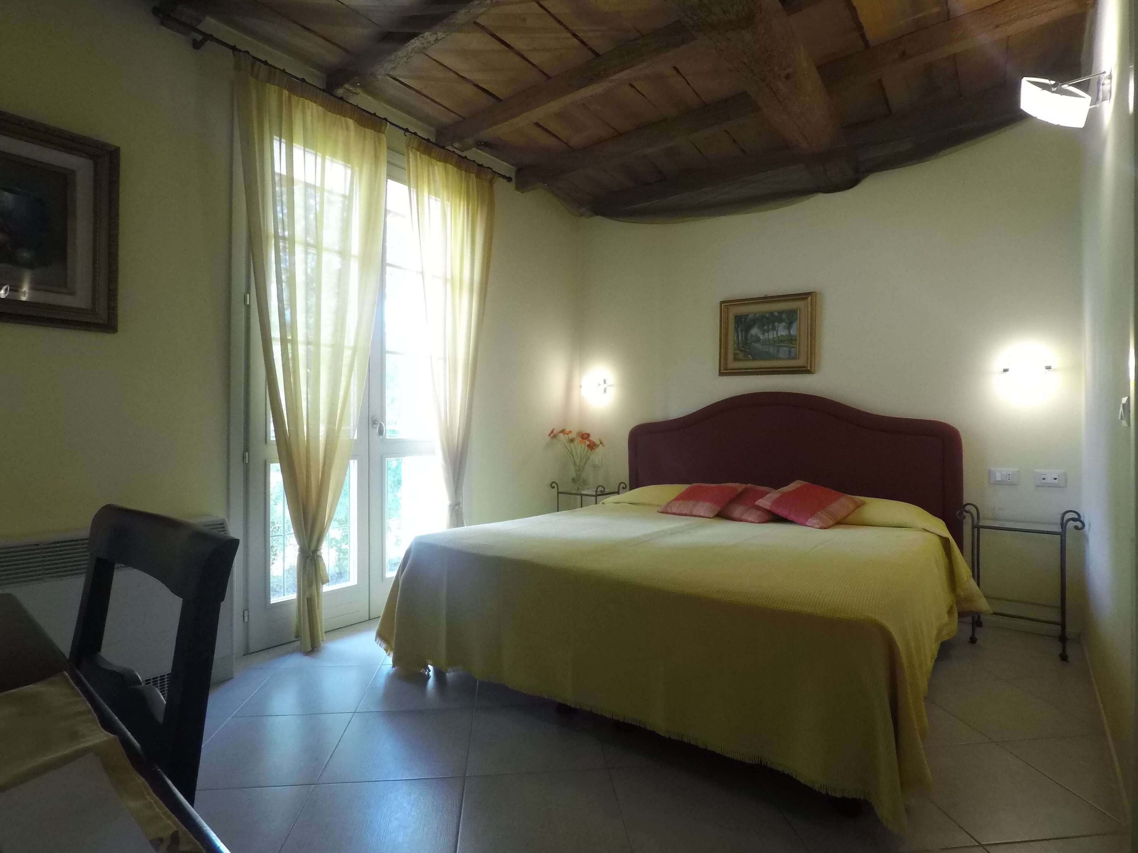 A bedroom with a bed, a chair, a window with curtains, a painting on the wall, and a ceiling light.