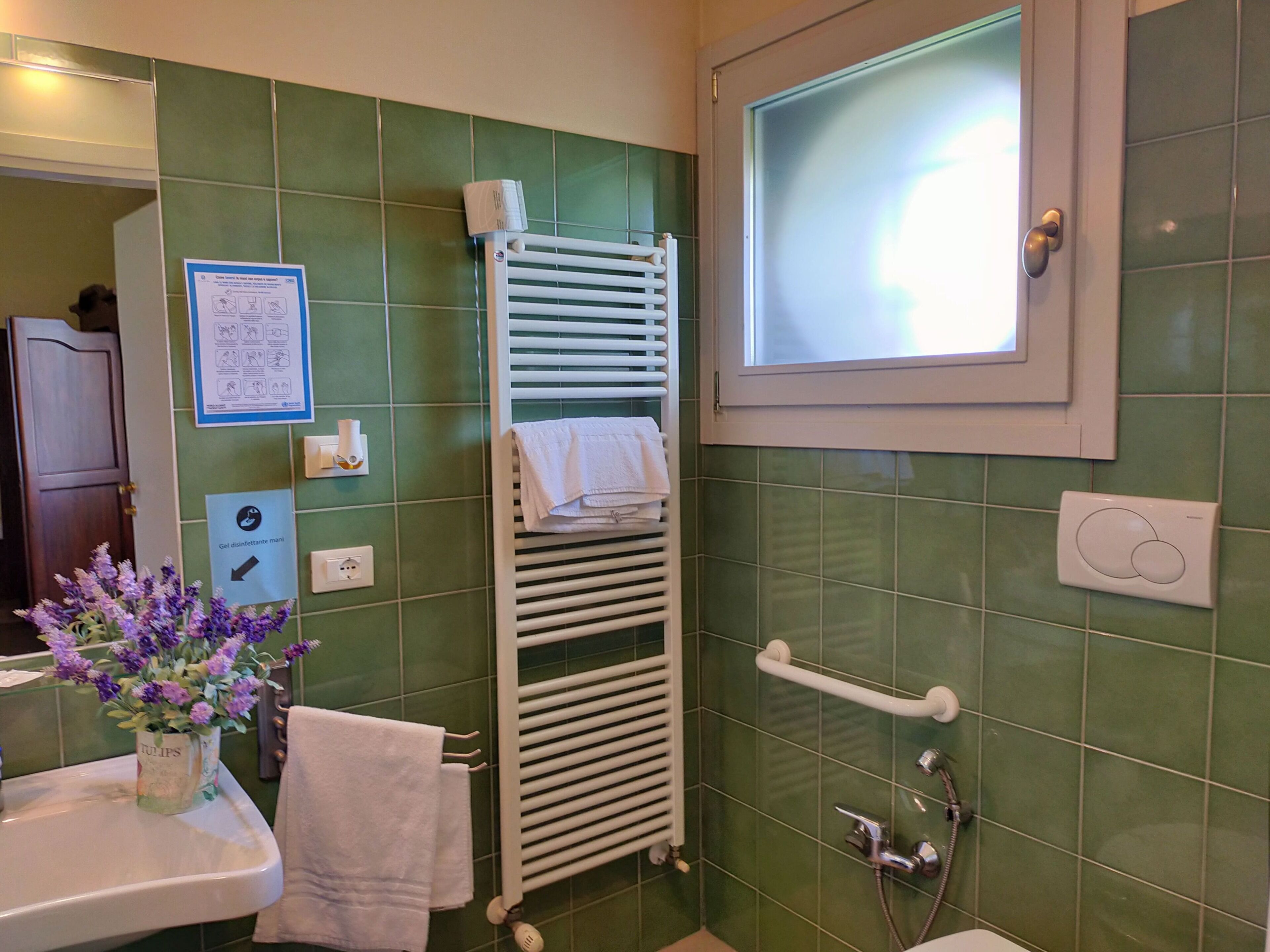 A bathroom with green tiles, a white towel rack, a window, a shower control panel, and a sign with instructions.
