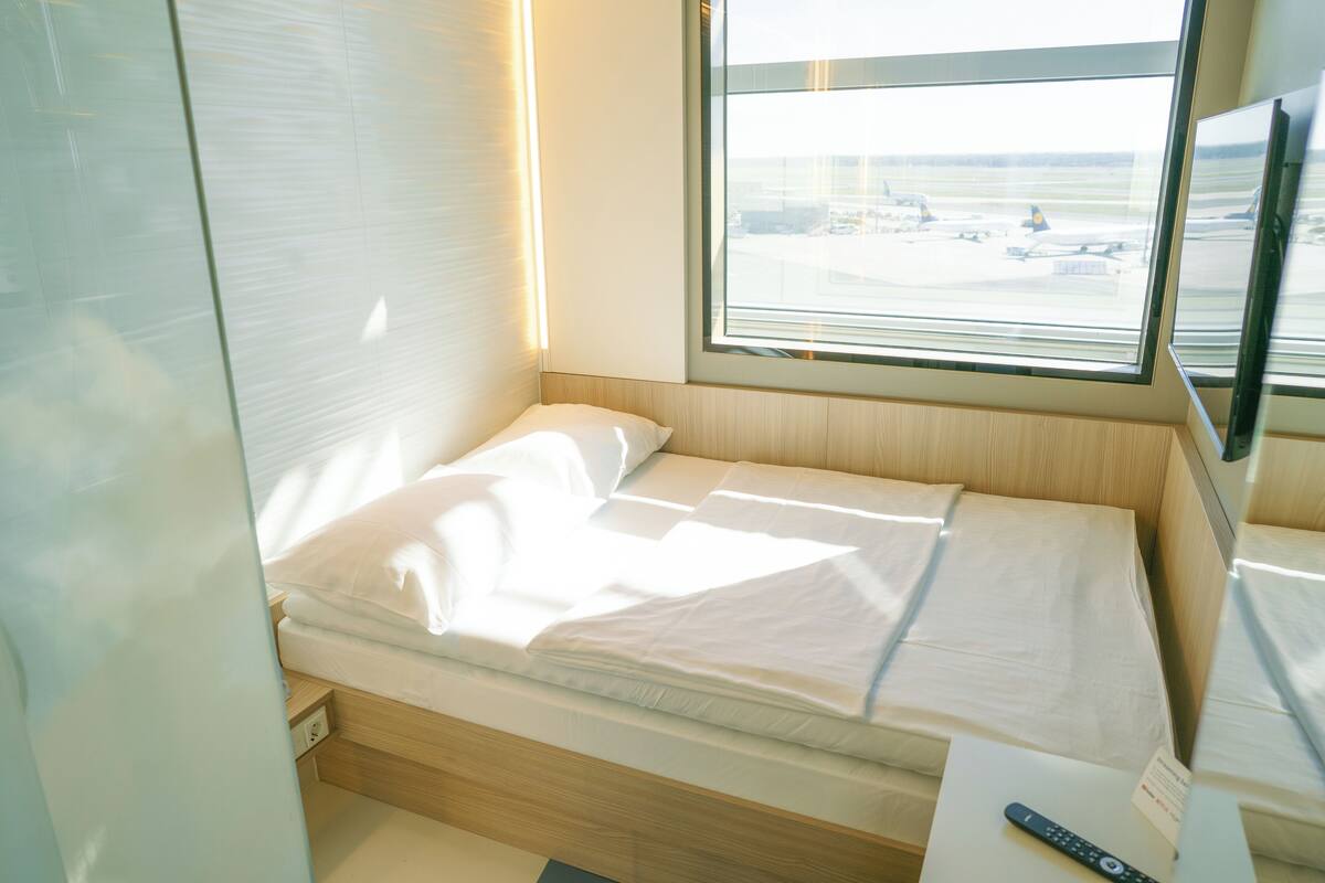 Ein Einzelbett mit Holz Kopfteil, ein Nachttisch und ein Fenster mit Blick auf einen Flughafen.