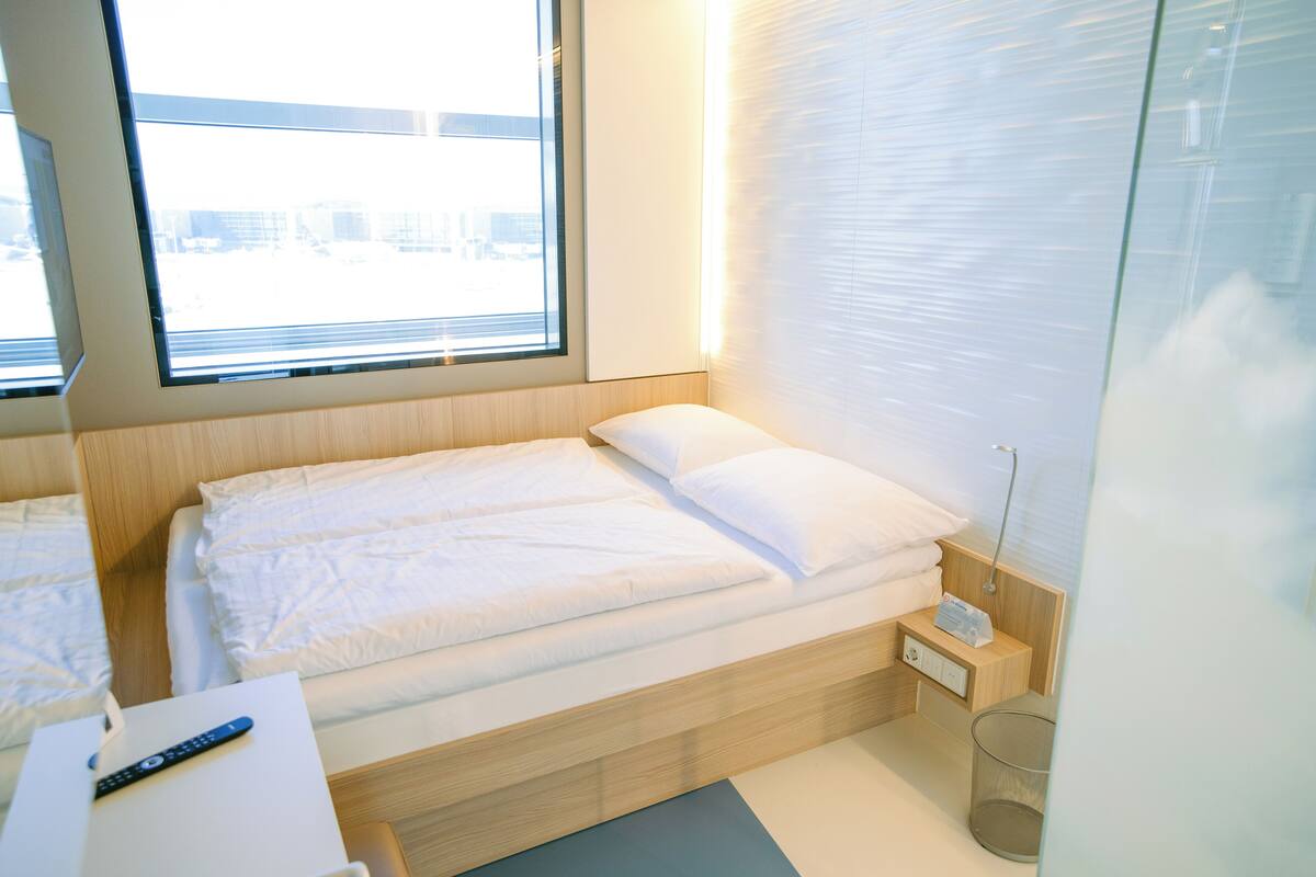 Ein modernes Hotelzimmer mit einem Holzbett, einem Nachttisch mit Fernbedienung und einem Fenster mit Jalousien.