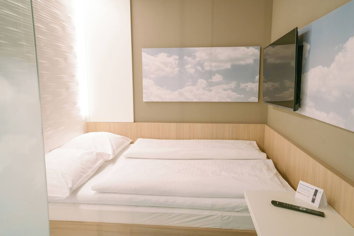 Ein ordentlich bezogenes Bett mit weißen Laken, eine Fernbedienung auf dem Nachttisch und ein an der Wand befestigtes Fernsehgerät darüber.