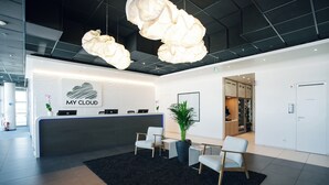 Un área de recepción moderna con un escritorio blanco, dos sillas blancas, un tapete negro y una planta grande en maceta.