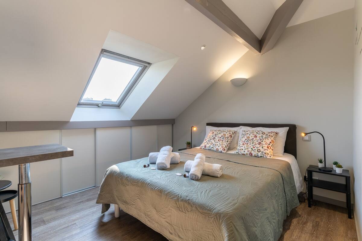 A modern bedroom with a skylight, a bed with floral pillows, and a bedside table with a lamp.