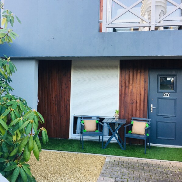 Un pequeño espacio al aire libre con dos sillas y una mesa, una puerta con un número y una ventana con una persiana cerrada.