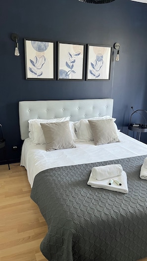 Cama bem arrumada com lençóis brancos e um edredom cinzento, mesa de cabeceira com abajur, e três quadros na parede.