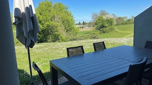 Um pátio externo com mesa e cadeiras, rodeado por uma área gramada com árvores e céu claro.