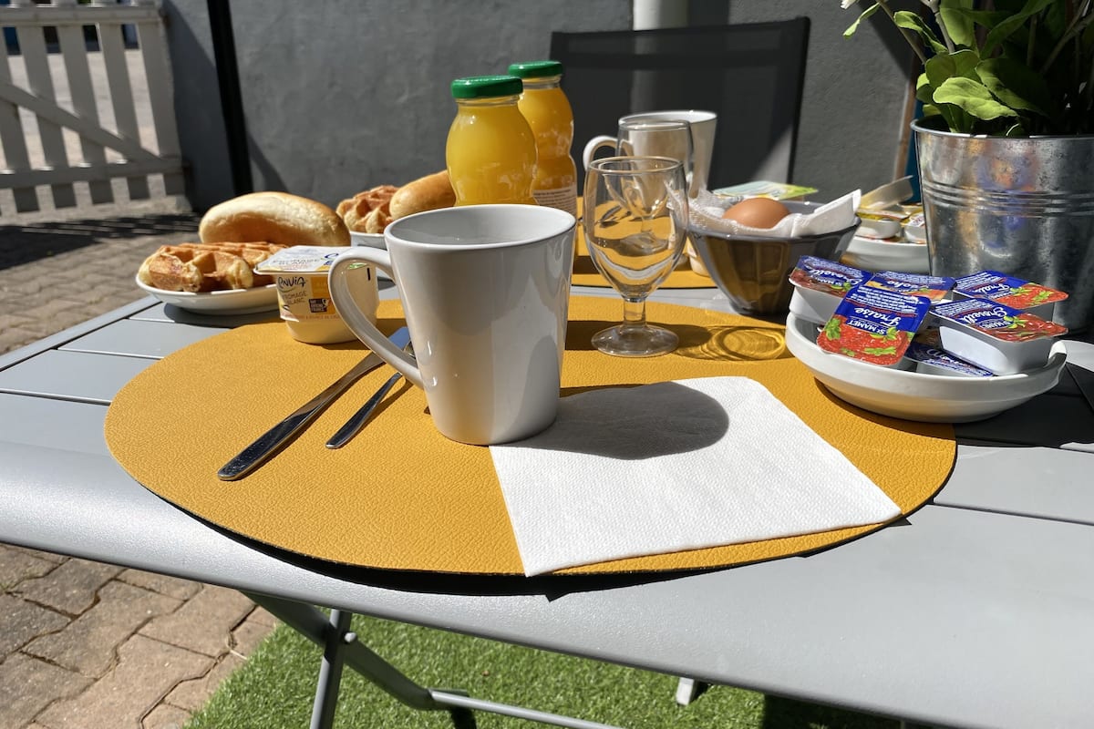 A breakfast table with a waffle, a mug, a glass, a bottle of juice, a bowl of yogurt, and a potted plant.