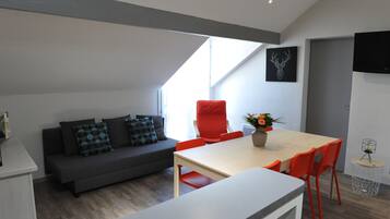 A modern living space with a sofa, dining table, and chairs, a TV mounted on the wall, and a ceiling with a skylight.