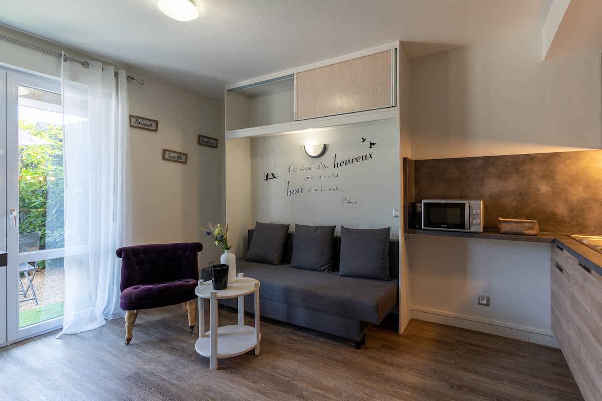 A modern living room with a grey sofa, a purple armchair, a small white table, and a wall with decorative text and a light fixture.