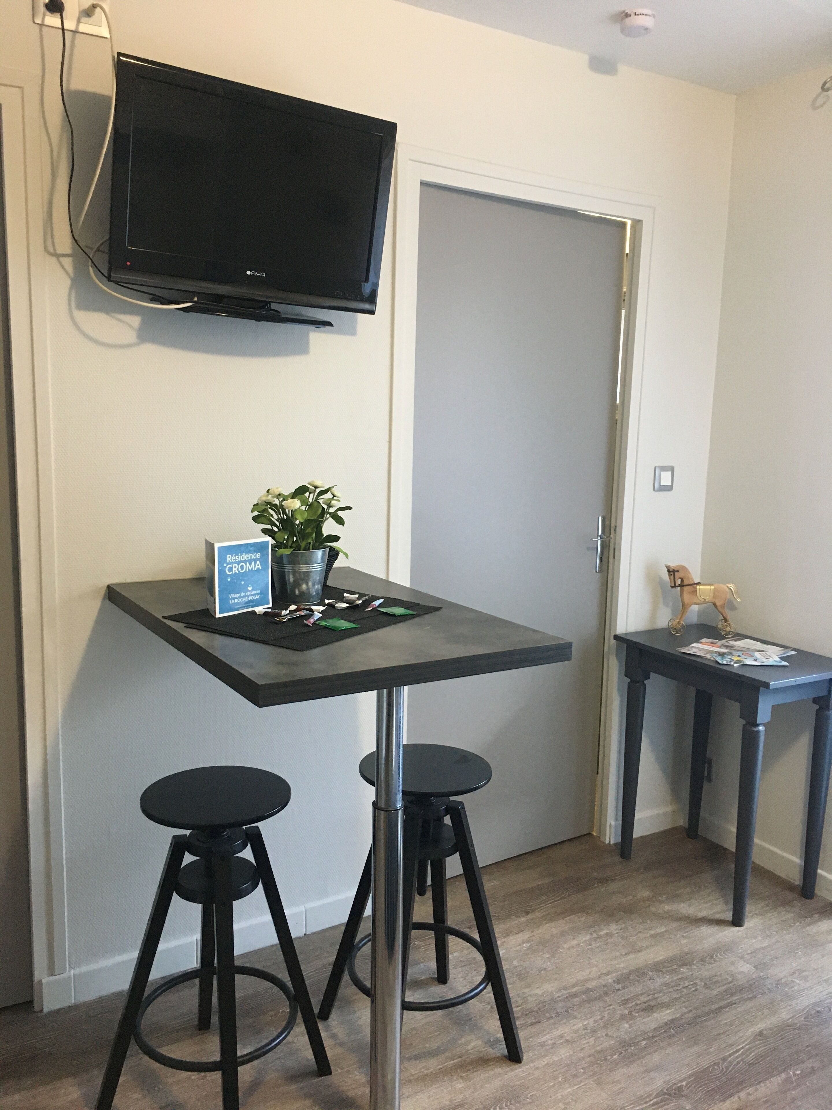 Una pequeña kitchenette con una televisión de pantalla plana montada sobre una encimera oscura, dos taburetes negros y una mesita con un caballito de juguete.