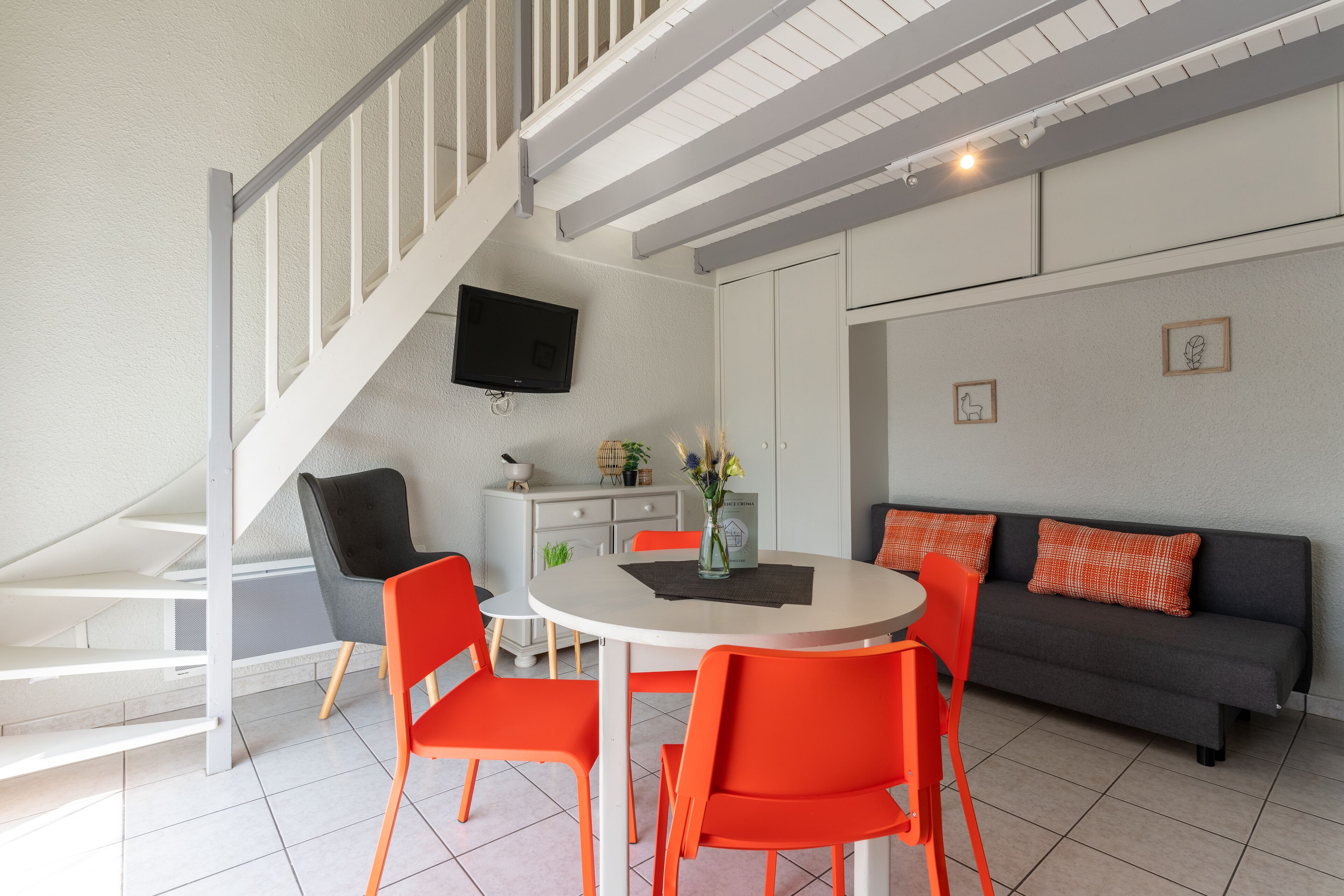 A modern living room with a staircase, a dining table with chairs, a sofa, and a flat-screen TV mounted on the wall.