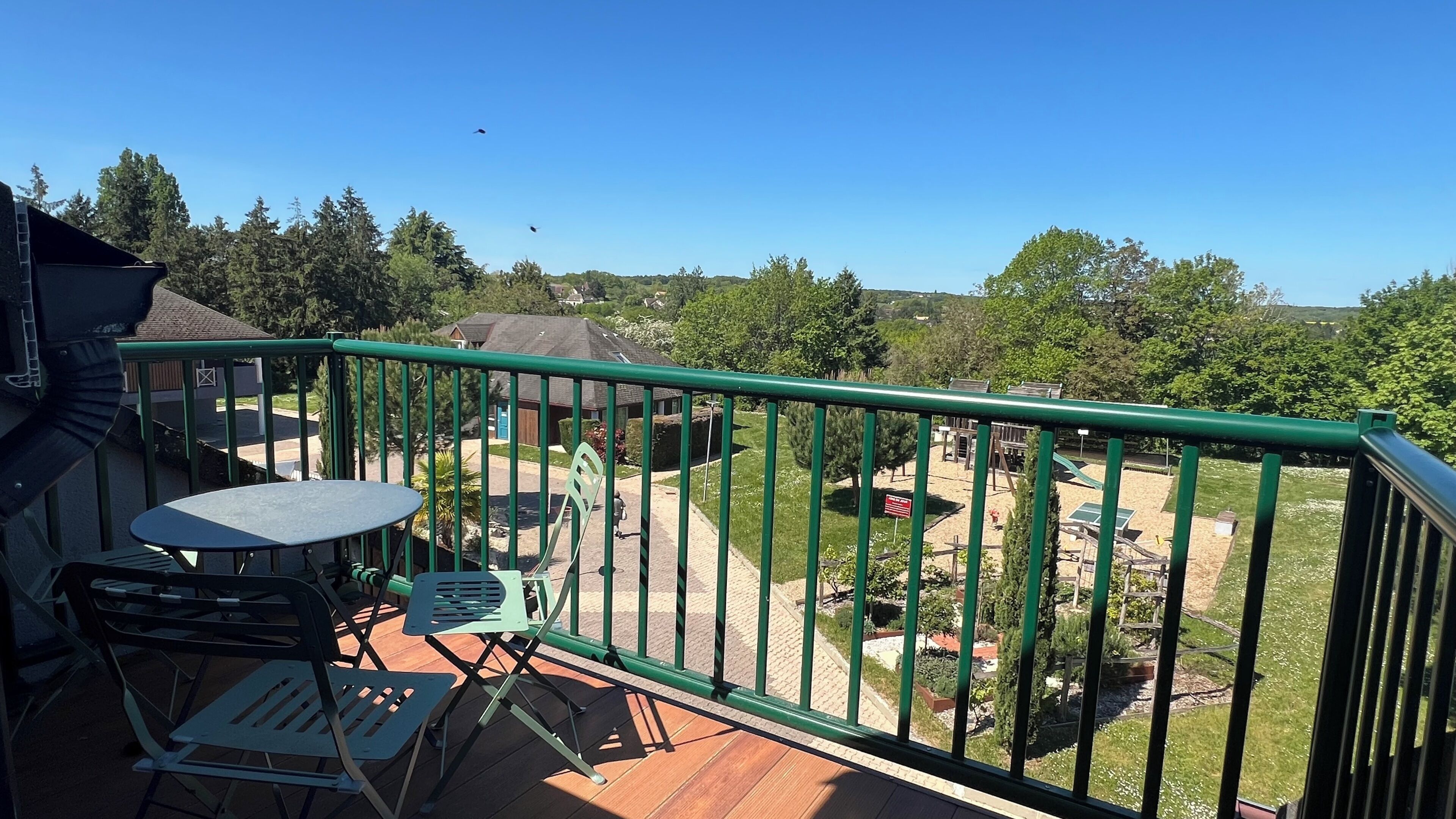 Un balcone con tavolo e sedie, vista su un'area giochi e cielo sereno.