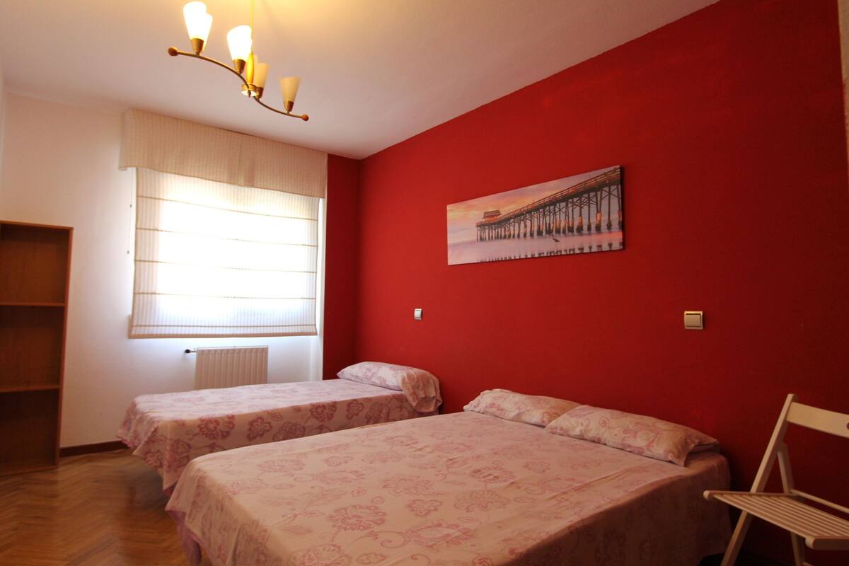 A room with two beds, a red wall, a window with blinds, a radiator, and a painting of a pier.