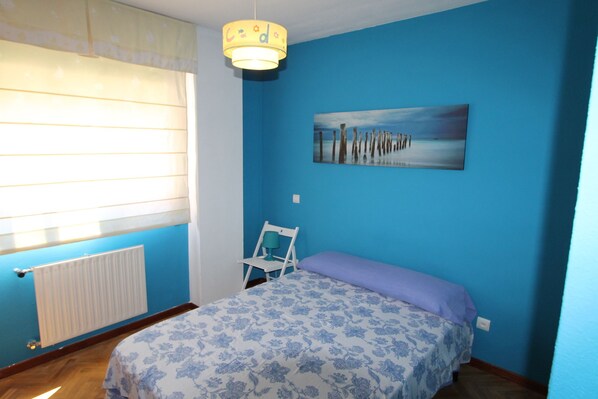 A bedroom with a bed, a chair, a radiator, a window with blinds, and a painting on the wall.