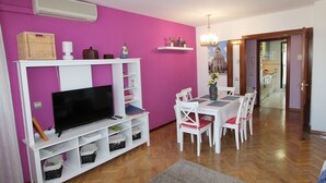 Ein Wohnzimmer mit einem Fernseher auf einer weißen Unterhaltungsecke, einem Esstisch mit Stühlen und einer leuchtend pinkfarbenen Wand.
