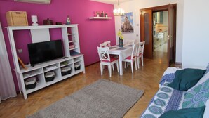 Ein Wohnzimmer mit Fernseher, weißen Regalen, einem Esstisch mit Stühlen, einer rosa Wand und Holzboden.