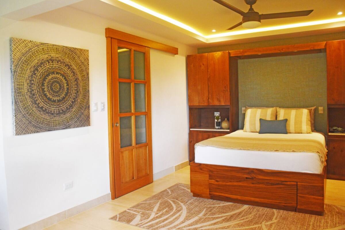 A bedroom with a wooden bed, a ceiling fan, a wall-mounted artwork, and a wooden door.