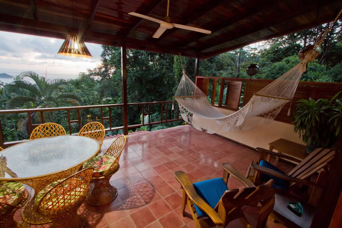 A covered outdoor seating area with a table and chairs, a hammock, and a view of the surrounding greenery.