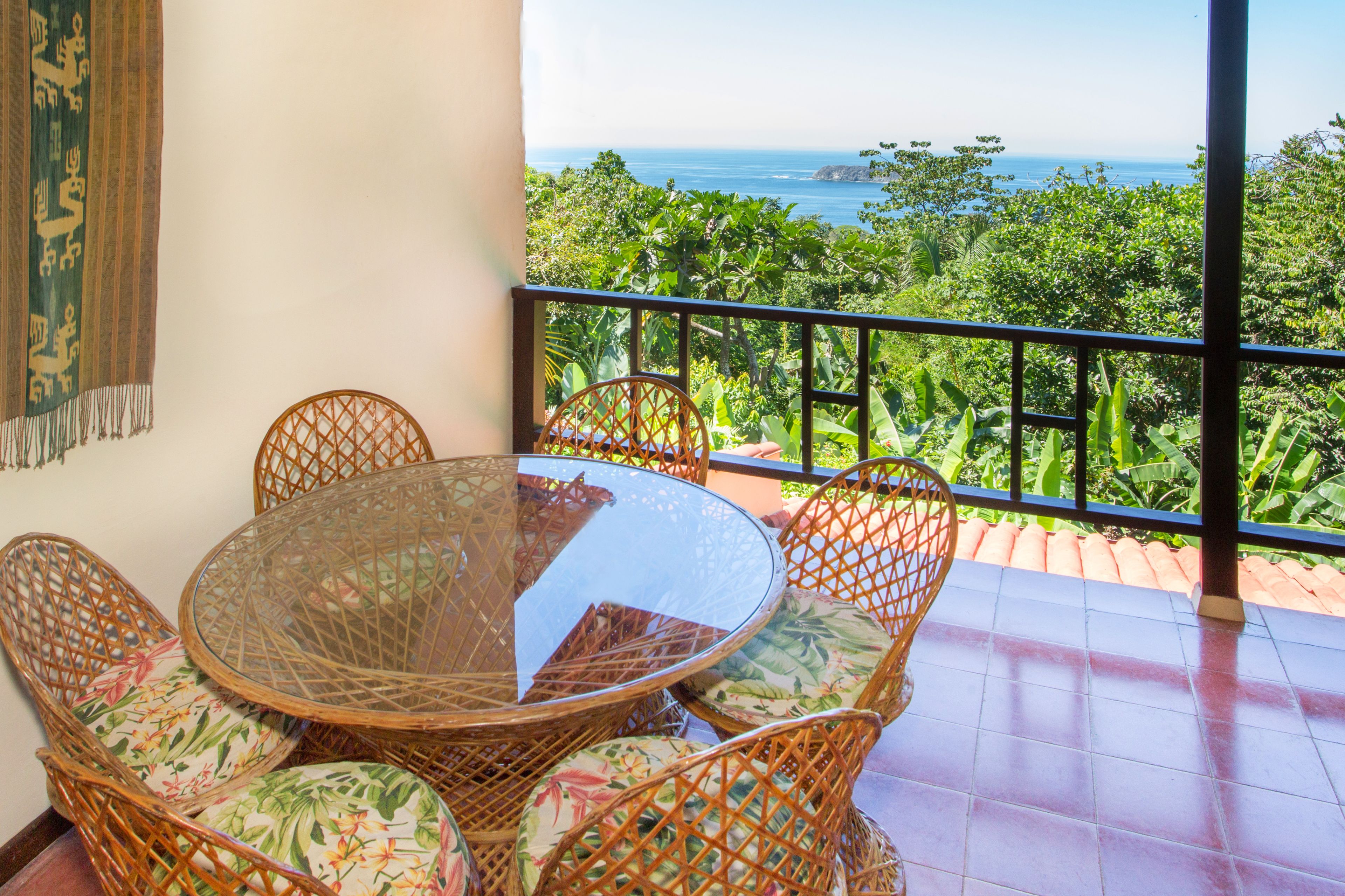 A round glass table with wicker chairs, a balcony with a view of the ocean, and a tiled floor.