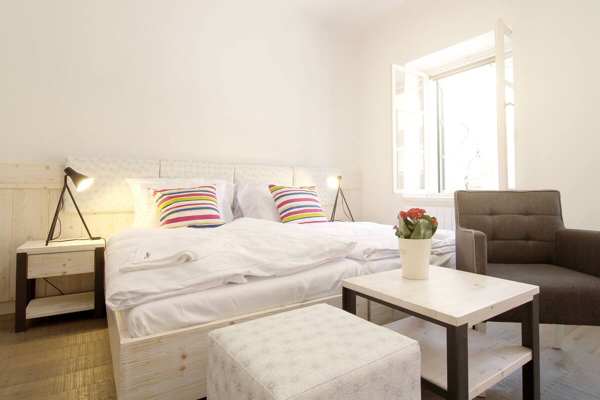 Une chambre à coucher avec un lit, des tables de chevet, une chaise, une petite table avec un vase et une fenêtre.