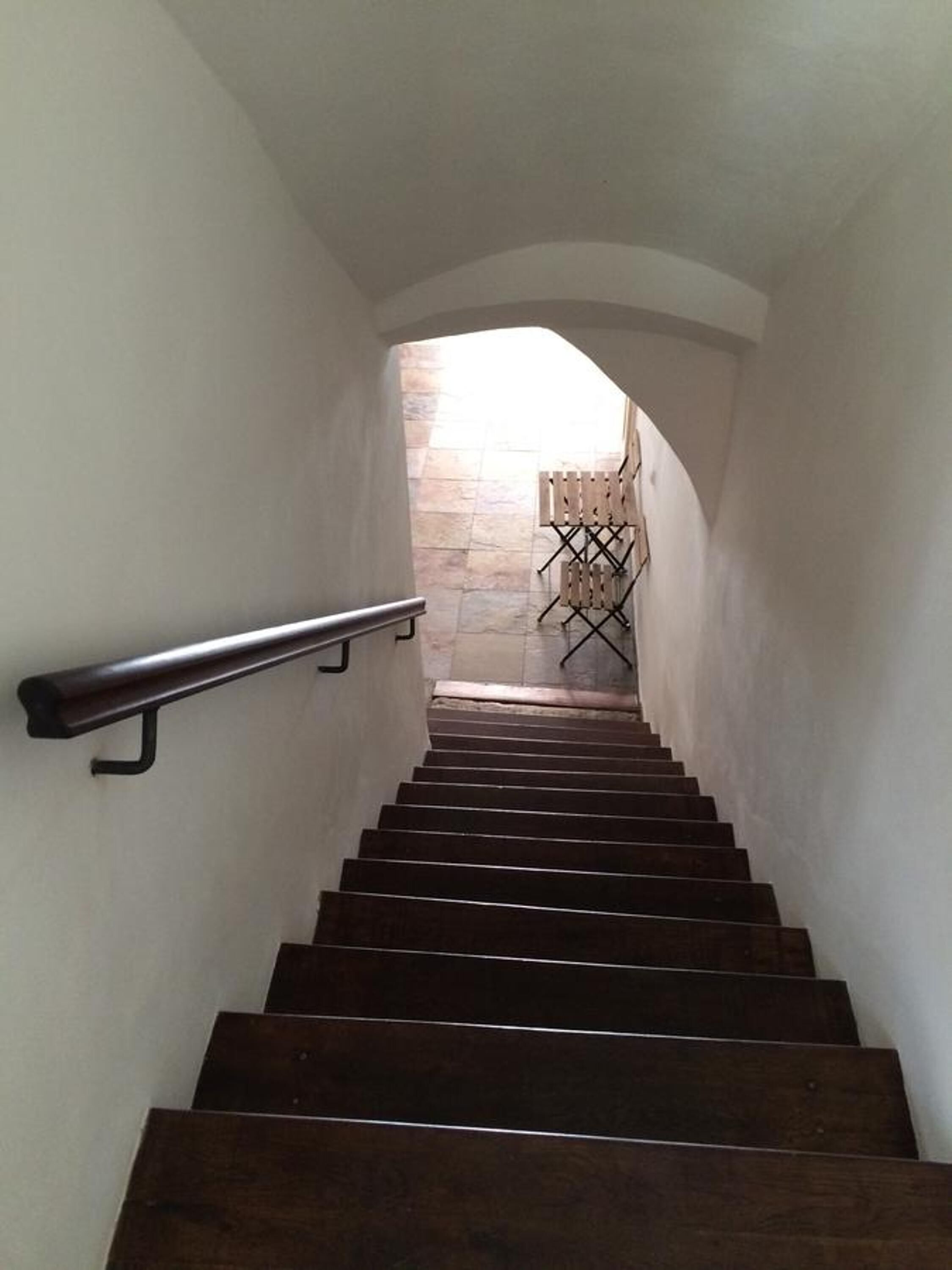 Una escalera de madera con barandilla metálica, que lleva a una pared de piedra y una silla plegable.