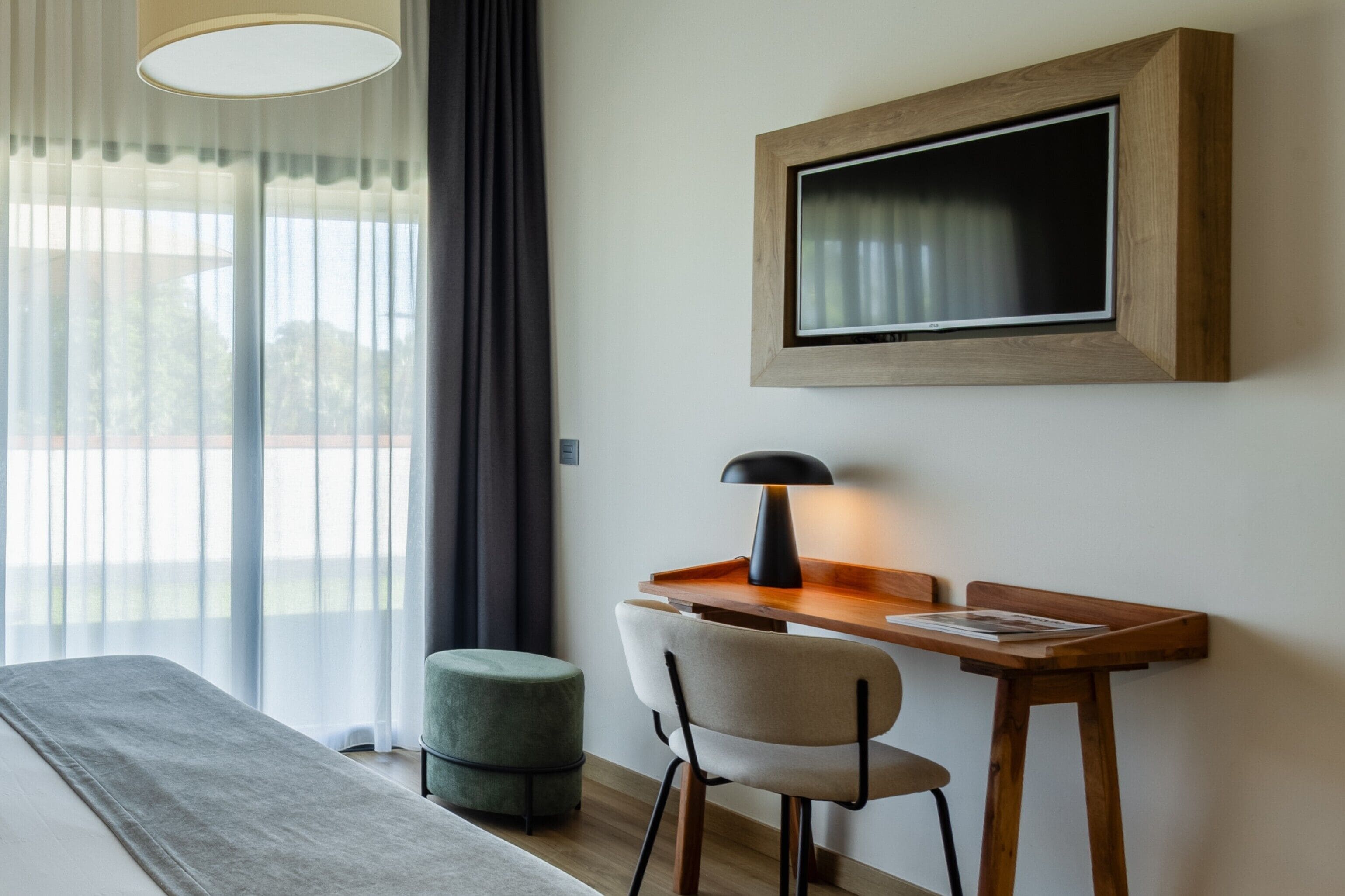 A bedroom with a bed, a desk with a chair, a small ottoman, a lamp, and a TV mounted on the wall.