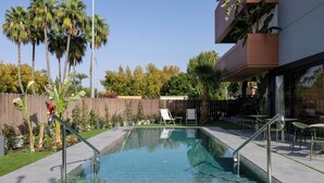 Una piscina rectangular con barandillas metálicas, rodeada por una cerca de madera, palmeras y un patio con mobiliario exterior.
