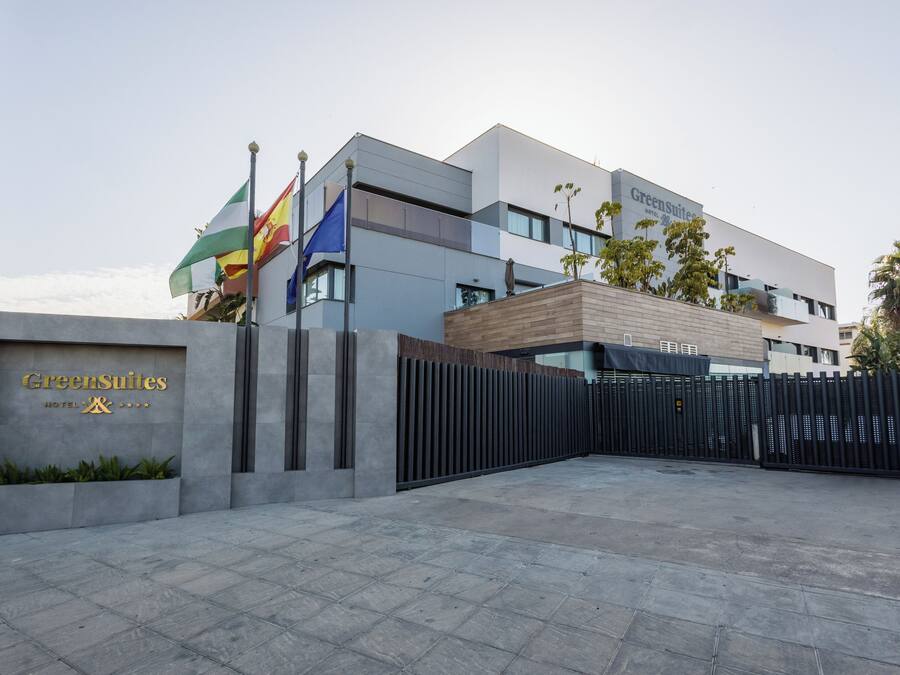 Una entrada de hotel moderna con un camino de acceso porticado, un cartel que dice 'GreenSuites Hotel' y varias banderas.