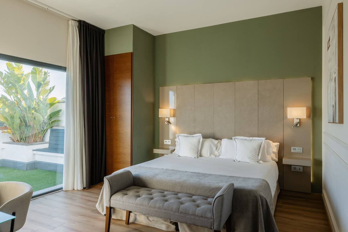 Une chambre d’hôtel moderne avec un grand lit, des lampes de chevet, un banc et une vue sur un jardin.