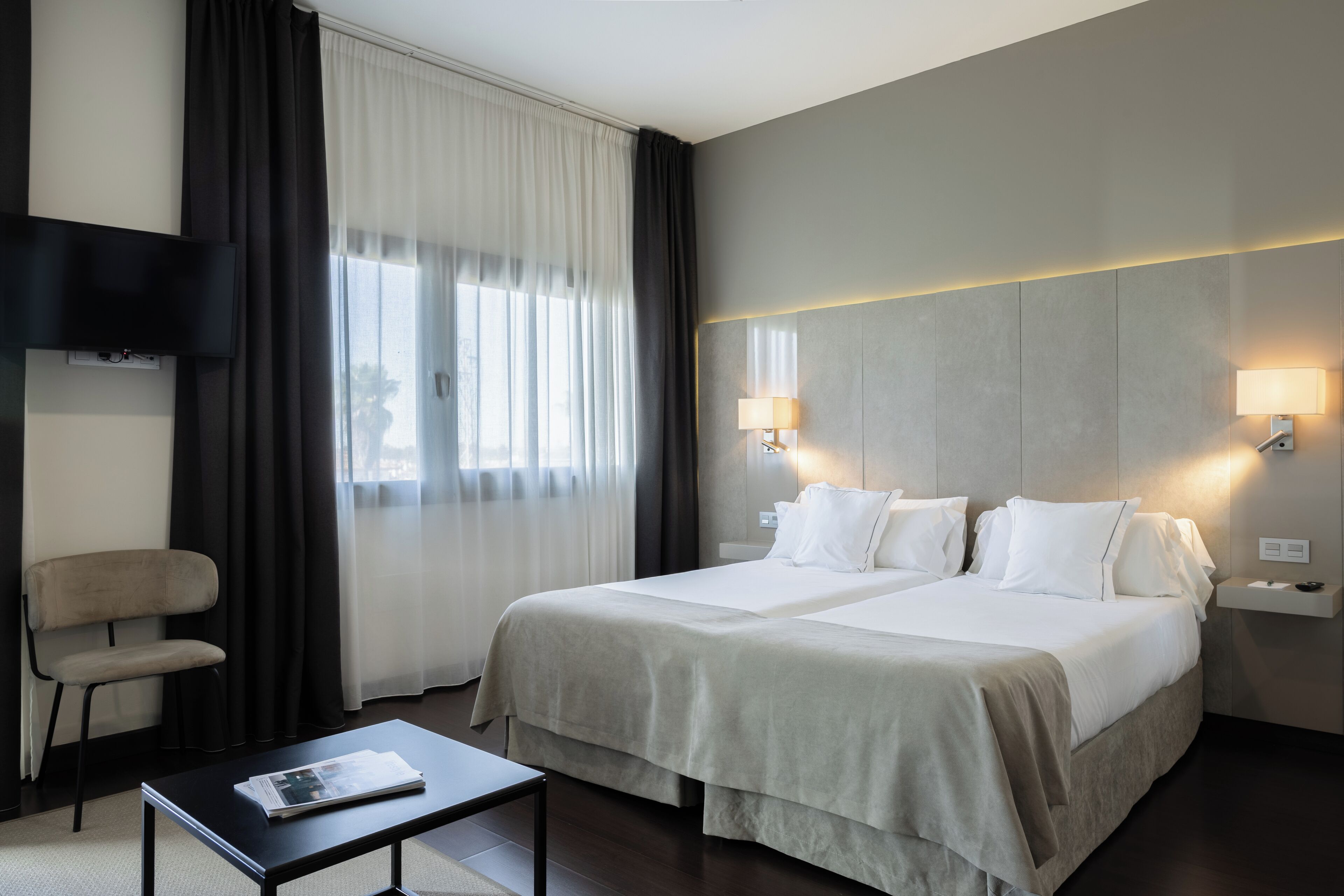 A hotel room with a bed, two bedside lamps, a TV mounted on the wall, a chair, and a table with a magazine.