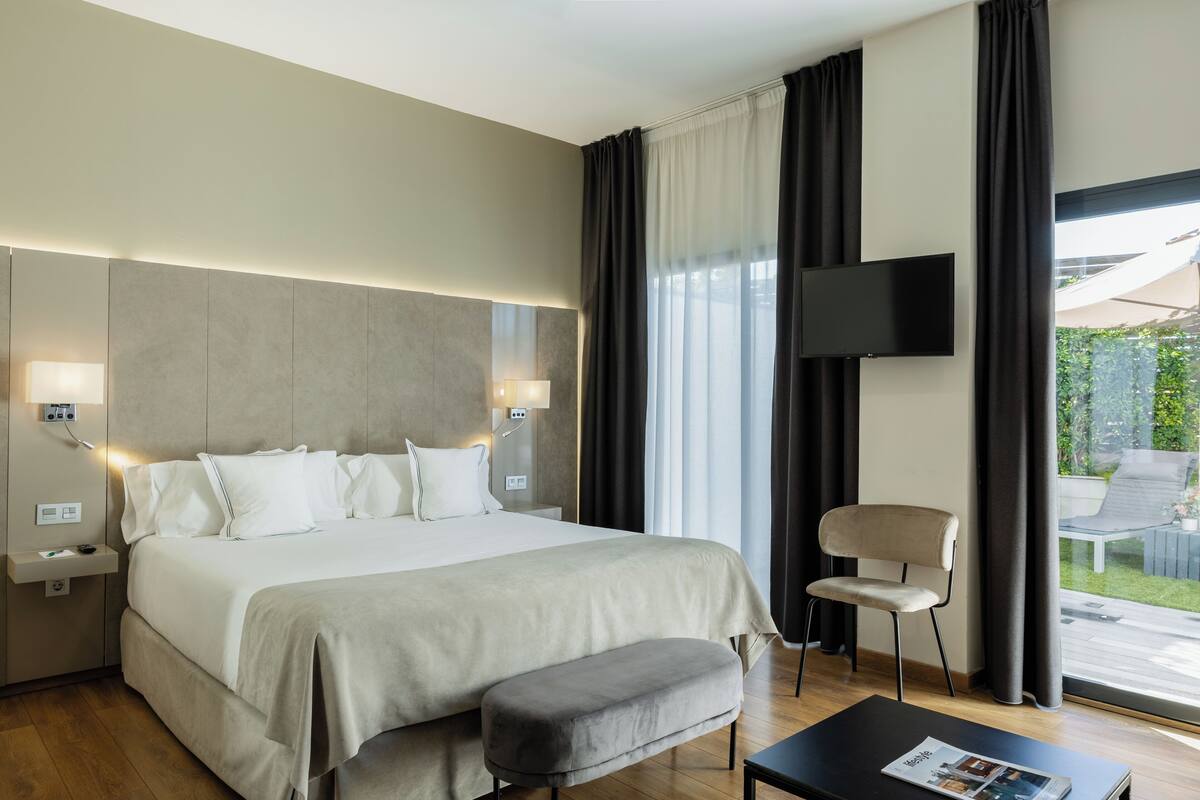 Une chambre d’hôtel moderne dotée d’un grand lit, de tables de chevet, d’un téléviseur à écran plat, d’un coin salon avec un fauteuil et d’une fenêtre avec des rideaux.