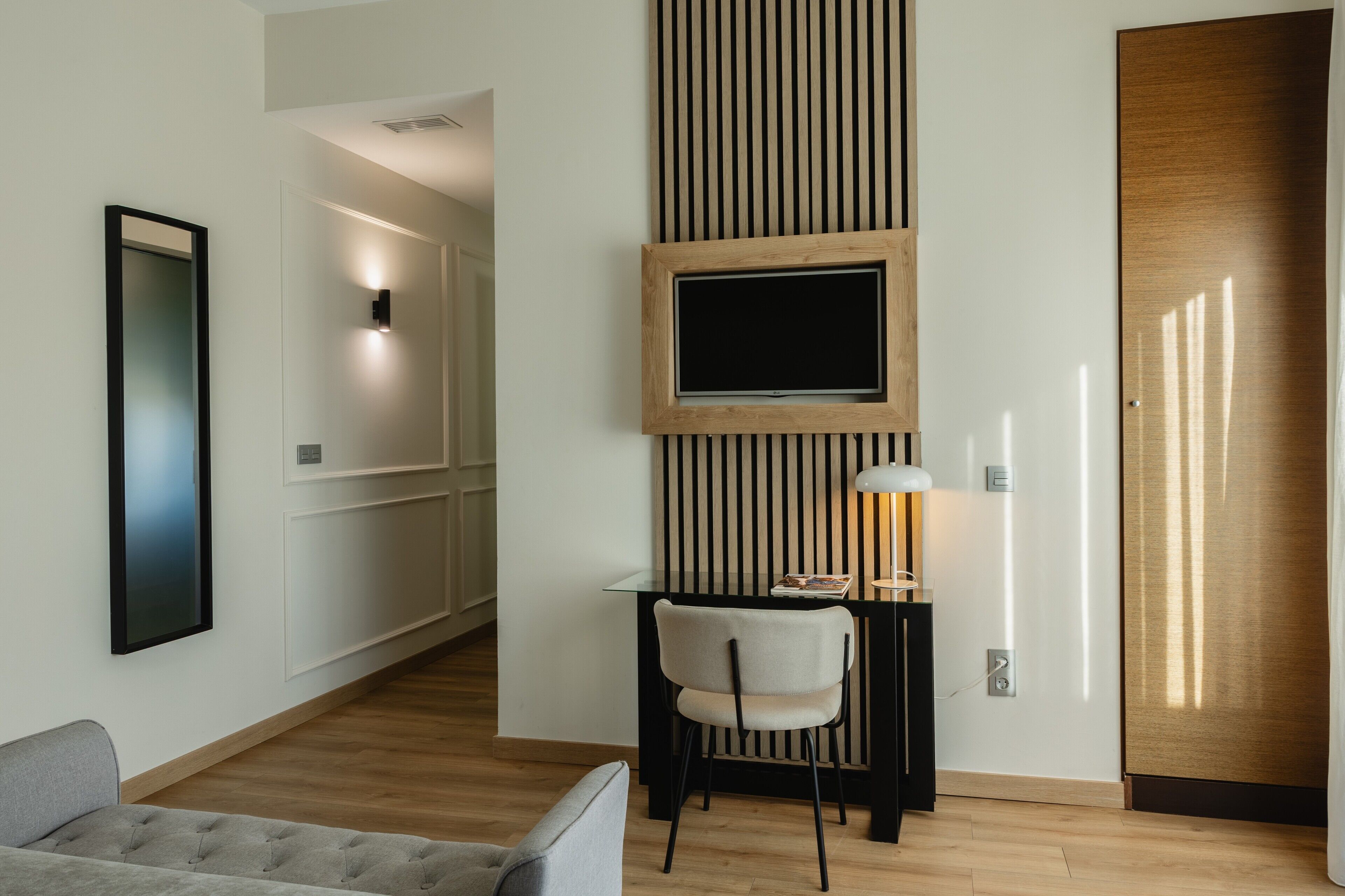 A modern hotel room with a wooden floor, a desk with a chair, a television mounted on the wall, and a door with a wooden panel.