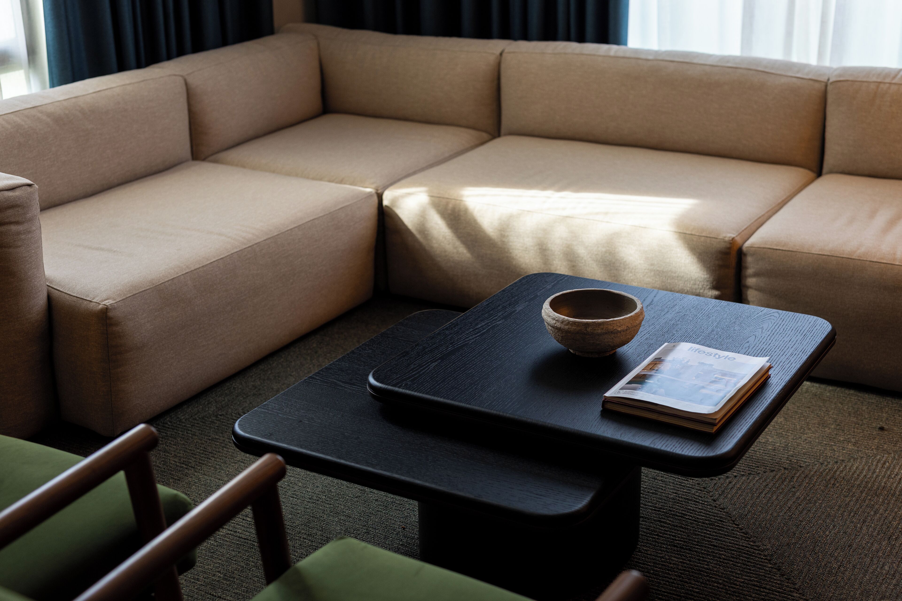 Un salon moderne avec un canapé d’angle beige, une table basse noire et un fauteuil vert.